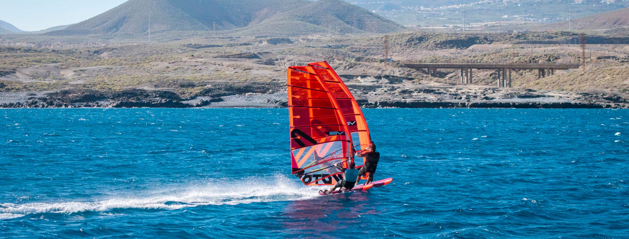 A windsurfer planing across blue water under an orange Severne sail, arid coastal hills behind